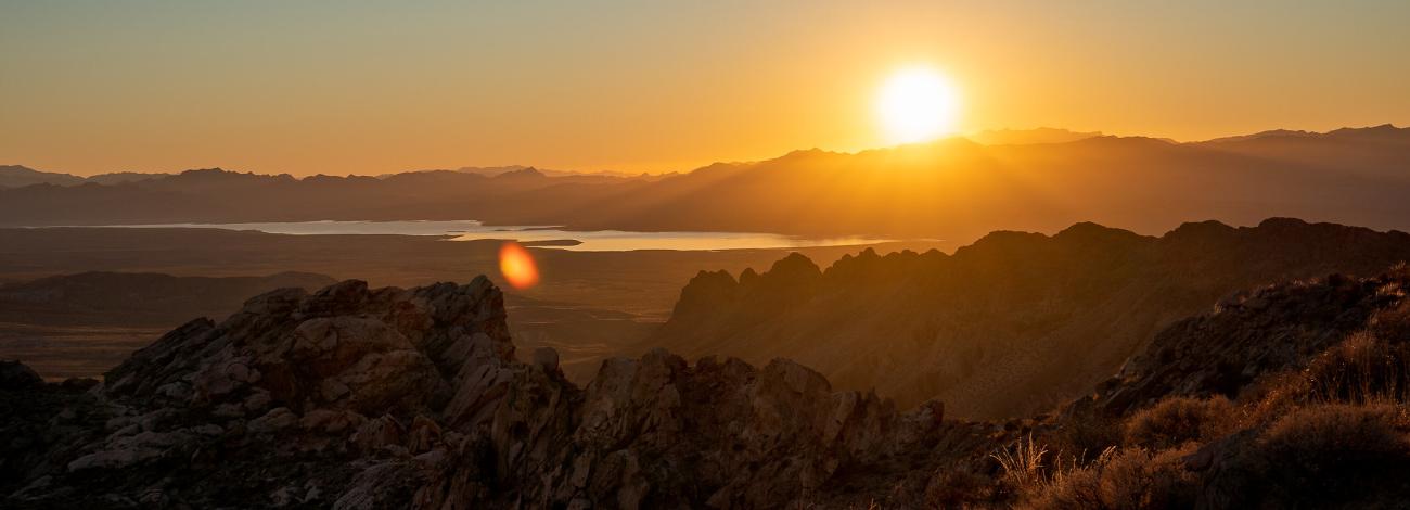 Sun rising just barely above the mountain range casting a bright orange and red light illuminating the valley, water and other mountains