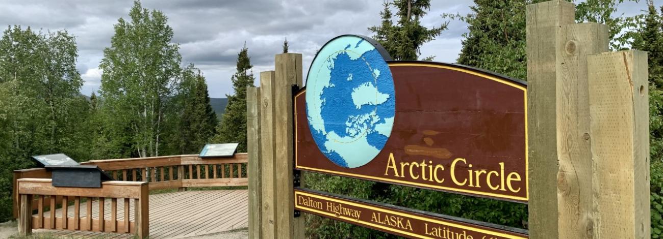A wooden sign for the Arctic Circle, Dalton Highway, Alaska, Latitude 66° 33’.  Depicts north pole of the earth with the arctic circle shown in a dashed line. 