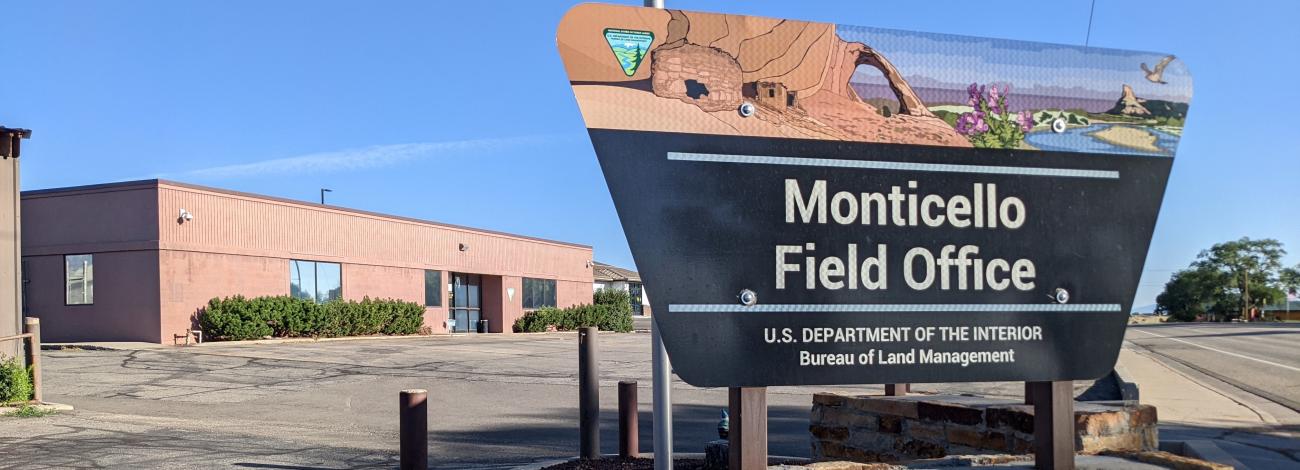 A colorful sign reading "Monticello Field Office" stands in front of a squat, light brown building.