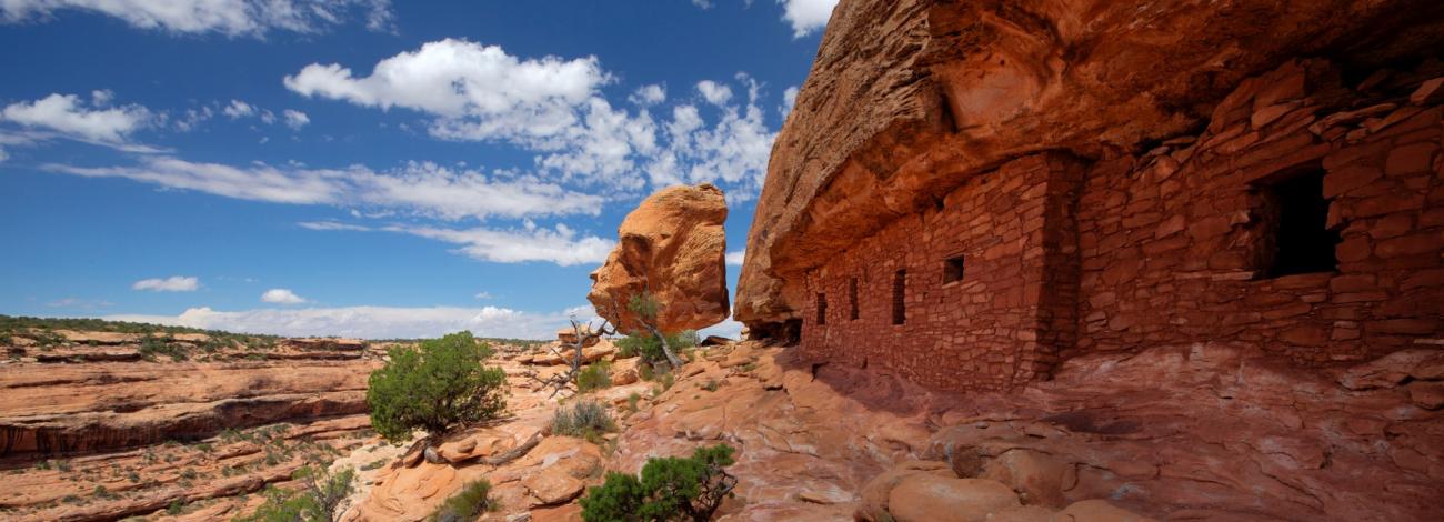 a cloudy blue sky shines brightly behind stone structures built into a redrock cliffside.