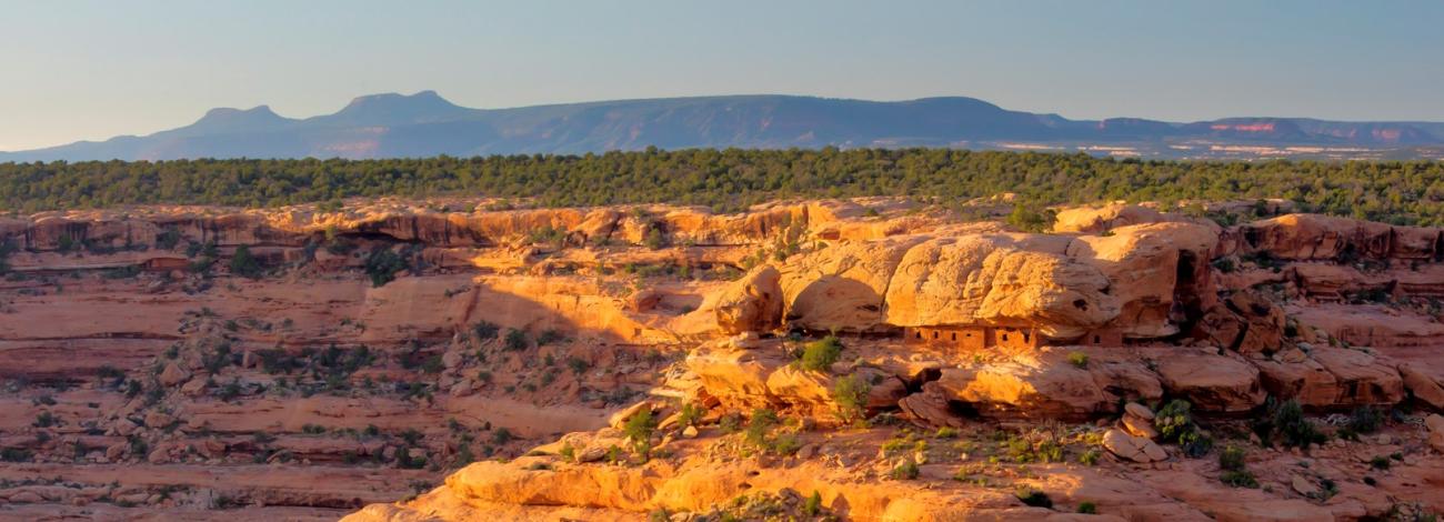 orange light illuminates a small archeological site nestled in an enormous cliffside. Buttes in the shape of bears ears loom in the distance.