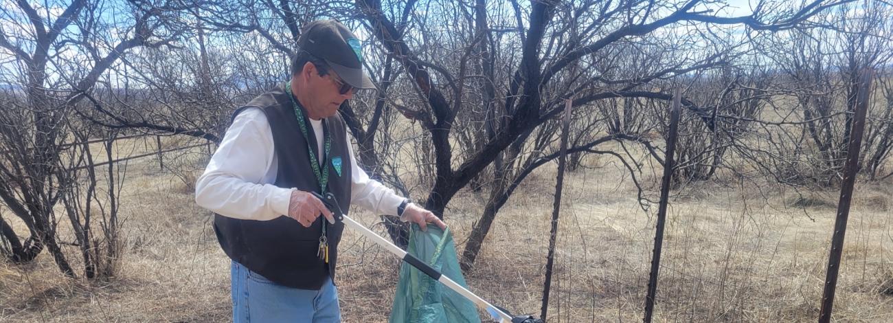 Man wearing a cap and vest with the BLM logo using a trash picker to remove a garbage bag full of trash from the ground