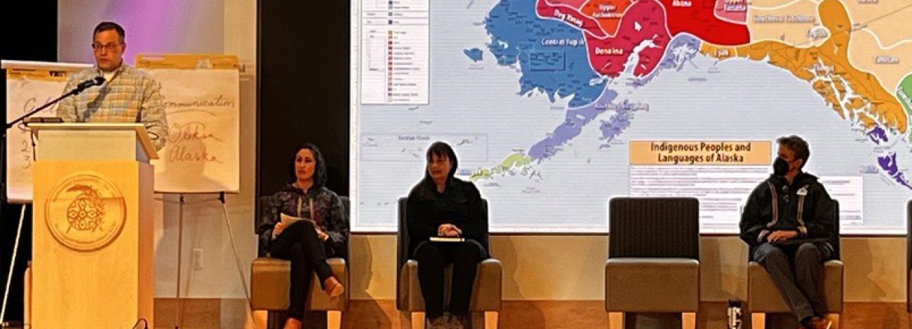 A person speaks at a wooden podium in front of a map of Alaska depicting Indigenous Peoples and Languages of Alaska.