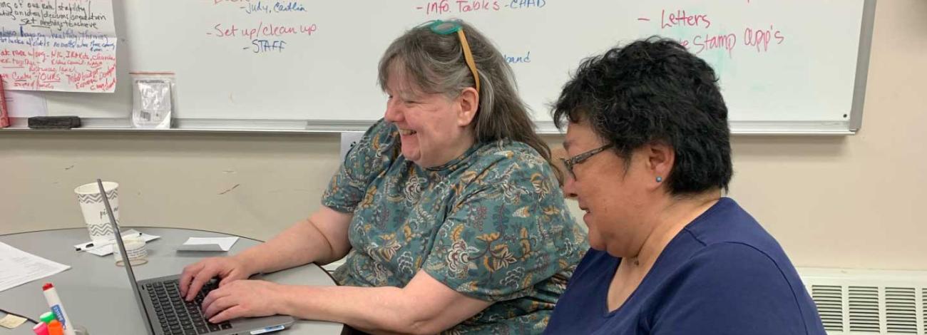 Two women sit smiling at a computer while they work to fill out an application.