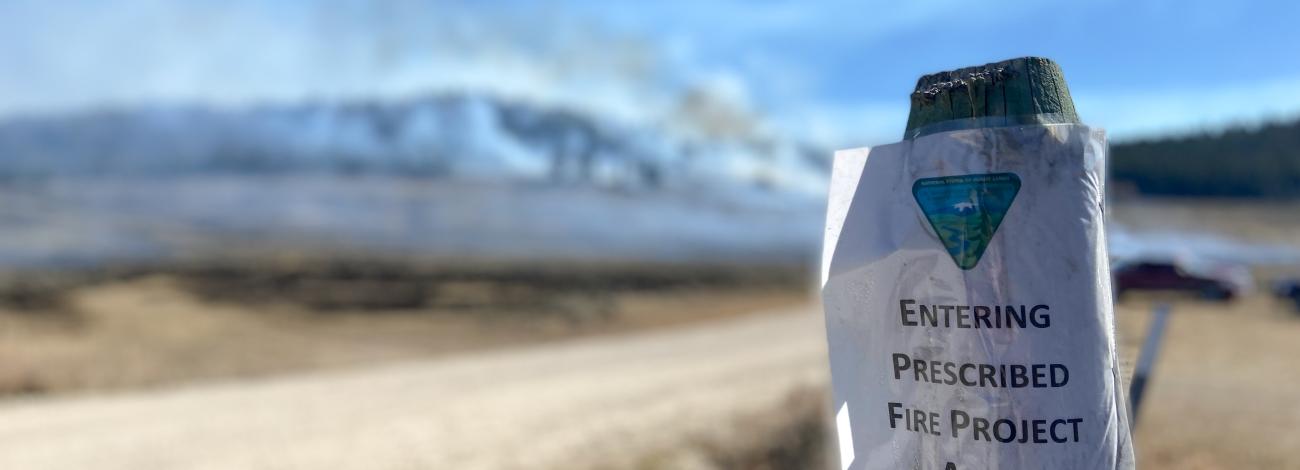 A sign on a fencepost reads prescribed burn area with smoke in background. 