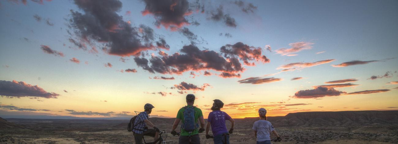 Four mountain bikers stop alongside a trail with their bikes to admire a colorful sunset lighting scattered clouds in pink hues.