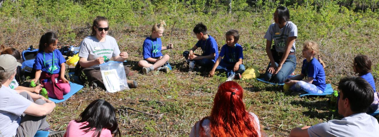Young students sitting in a circle gather around an instructor for outdoor learning experiences.