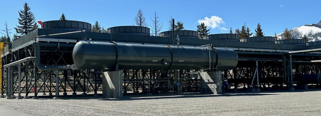 A landscape image of the Casa Diablo IV geothermal plant consisting of turbines, various steel pipes and tubes. A snowy mountain in the background.