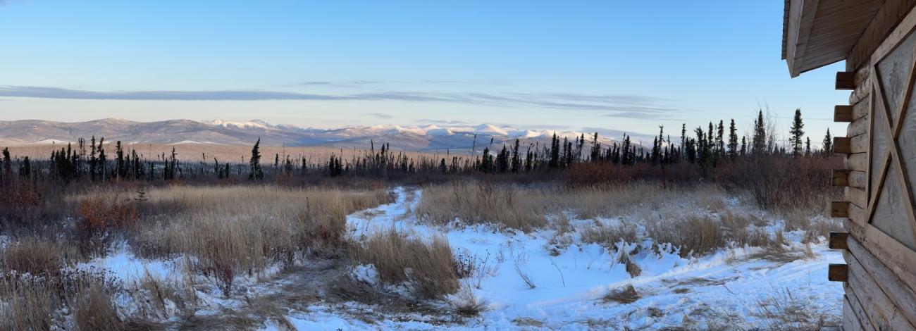 Picture of the view of the valley with the sun shining on the White Mountains' highlands in the distance. A few inches of snow covers the ground. The edge of the Moose Mountain Cabin frames the right side of the picuture.