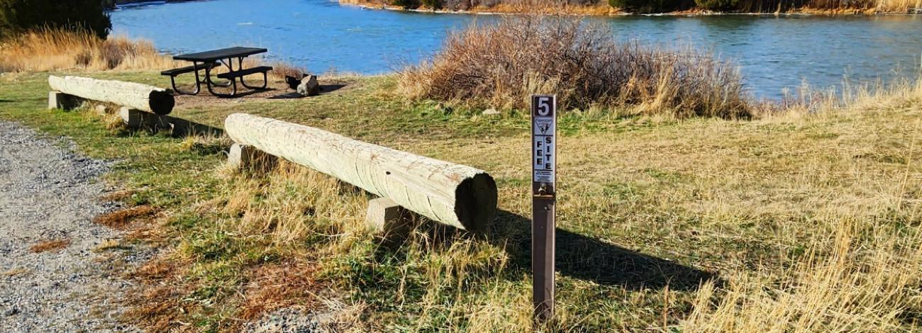 Log benches and a metal picnic table sit on the bank of a river.