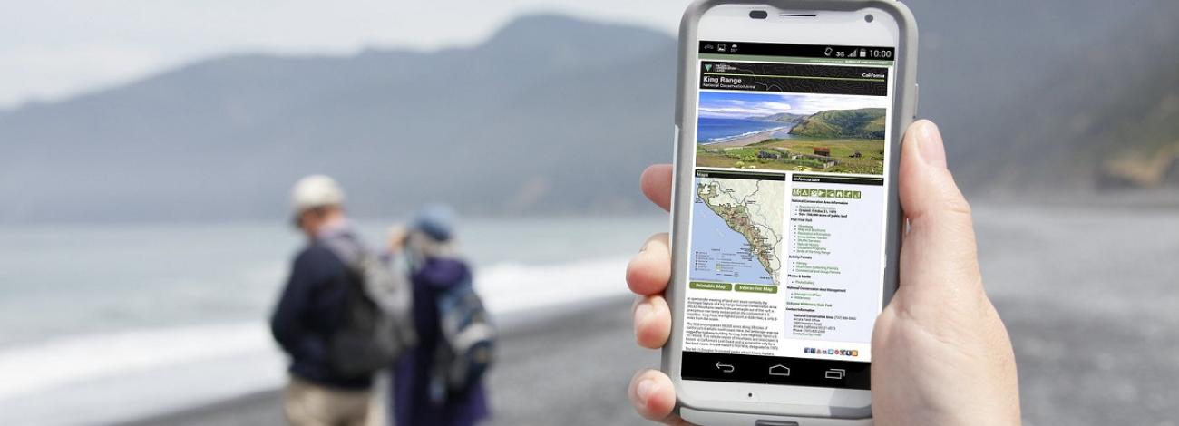 Hand holding cellphone with BLM web page of California's King Range on the screen while two people stand on beach facing water.