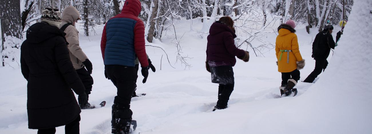 A group of people wearing winter gear snowshoe through deep snow.