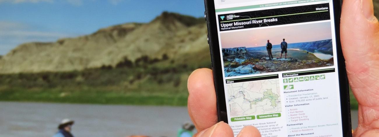 Hand holding cellphone with BLM web page of Montana's Upper Missouri River Breaks on the screen while two people are in a canoe on the water..