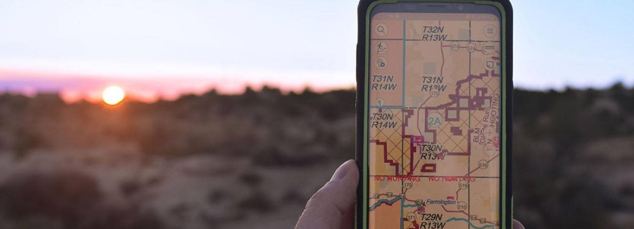 Hand holding cellphone with map of Public Land Survey System in Farmington New Mexico area on the screen and sun on horizon.