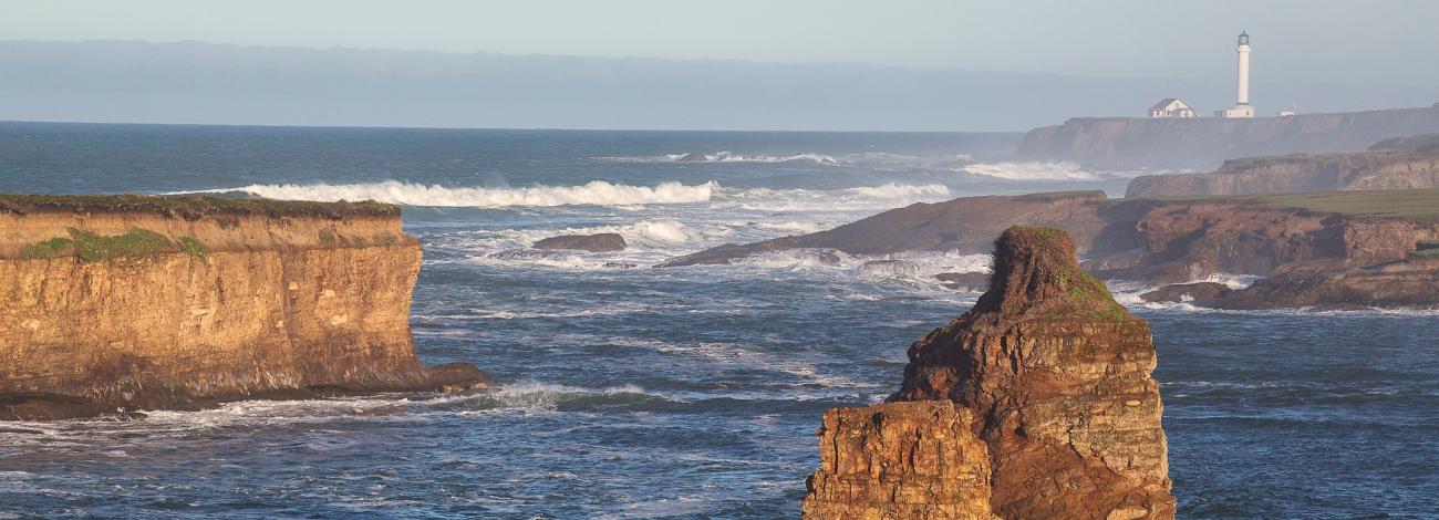 Ocean wave crash against a bluff as a light house shines bright.