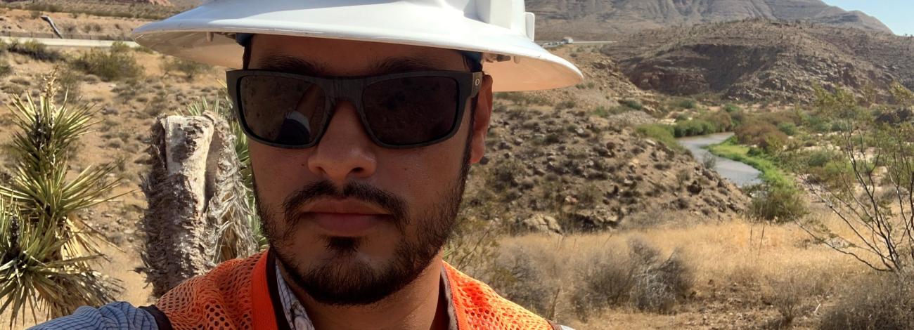 Carlos Joya, BLM Arizona's state engineer, takes a selfie. He is wearing a white hardhat, black glasses, and an orange high-visibility vest. The Virgin River Campground area is behind him.