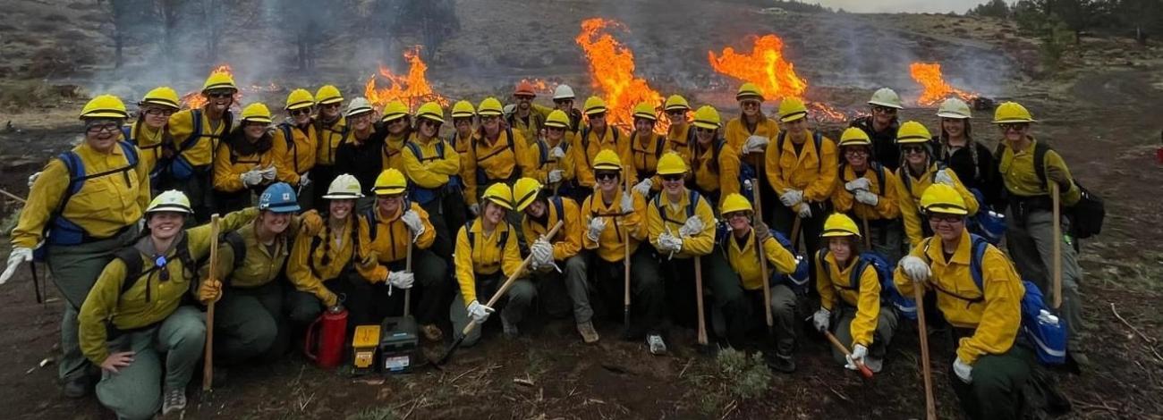 group shot of several dozen firefighters wearing yellow shirts and hardhatsposing in front of a fireline