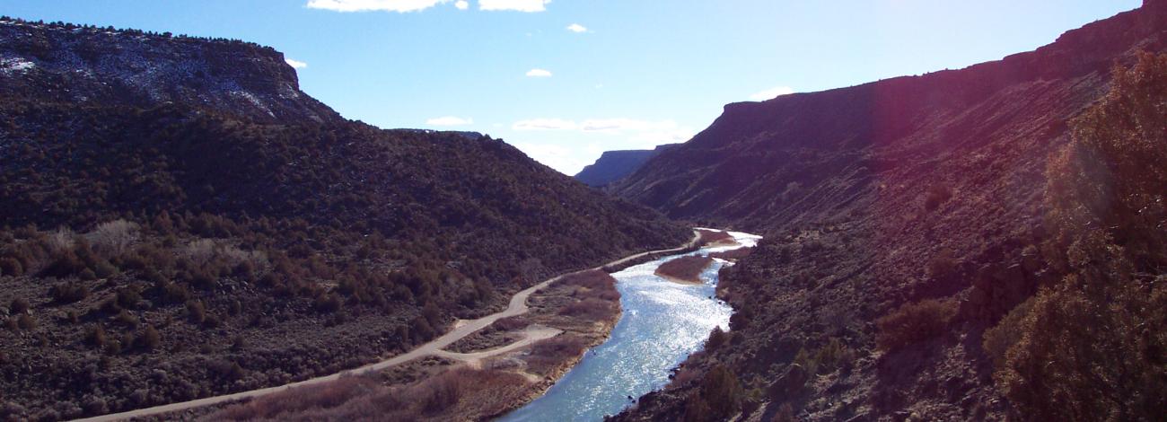 A wide photo of a river running through a deep brown canyon, with blue skies and bright sun in the background.