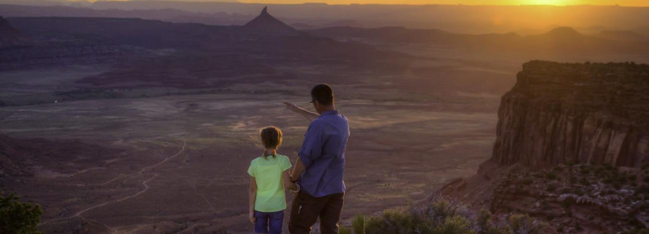 Photo shows a man with a blue shirt and brown pants holding a child’s hand, while standing on the edge of a canyon overlooking a desert valley landscape. The child is wearing a lime green shirt and blue shorts. The sun is setting in the background. 
