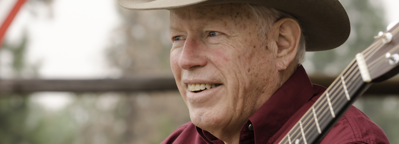 A man with a cowboy hat wearing a red shirt holds a guitar. 