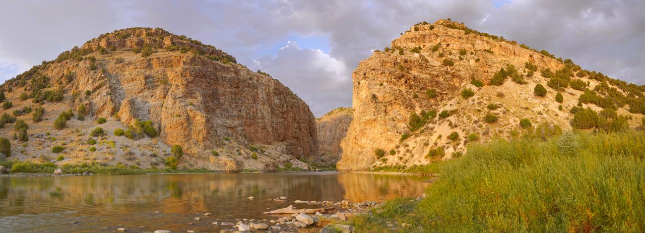 A serene landscape featuring two rocky cliffs surrounding a calm river. The cliffs are sunlit with patches of vegetation, while green reeds line the water's edge. Partly cloudy skies complete the natural scene.