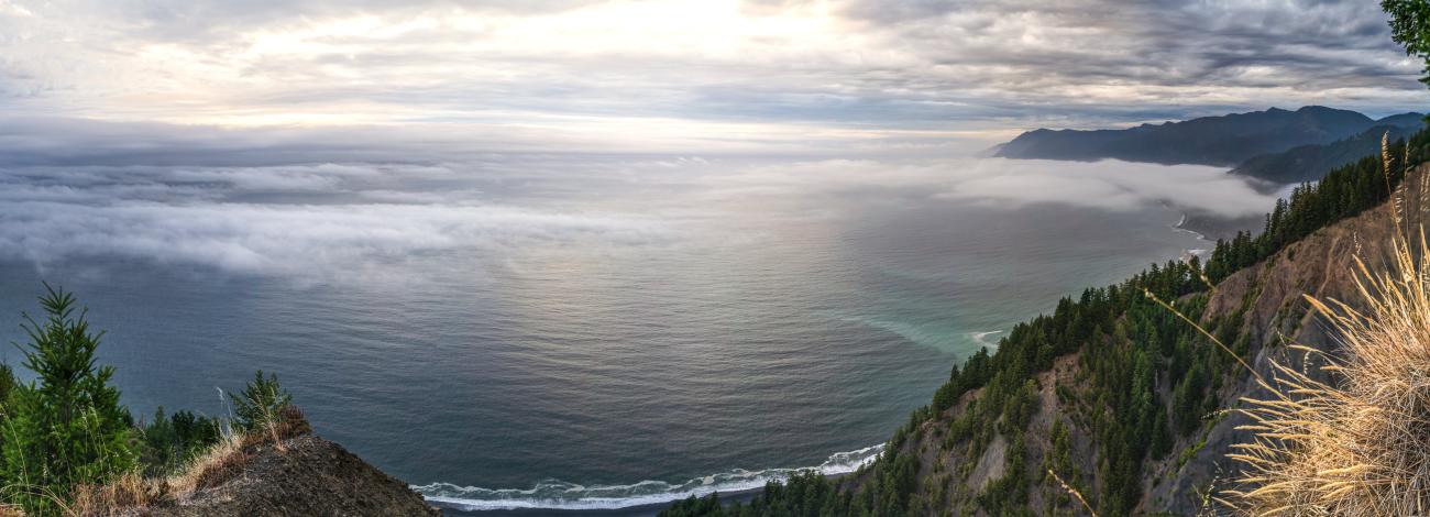 A wide view of the Pacific Ocean at sunset. Pine trees and tall, forested, mountain ranges frame the view.