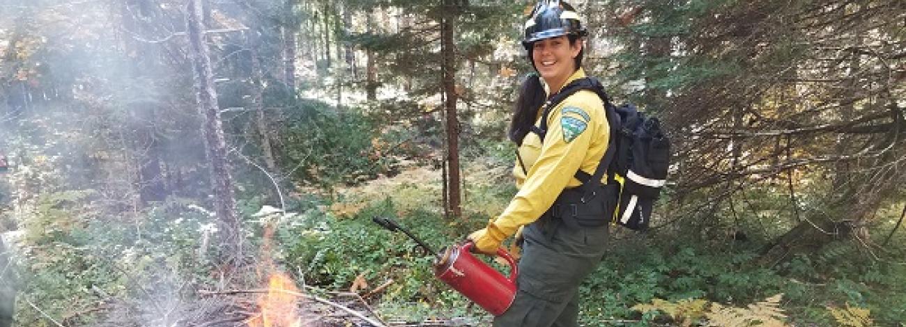 smiling firefighter in traditional yellow shirt holding a drip torch near a burn pile