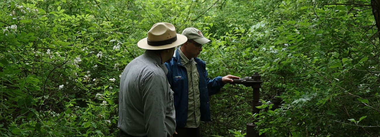 2 men stand next to a wellhead in a forested area, one rests his hand on it. 