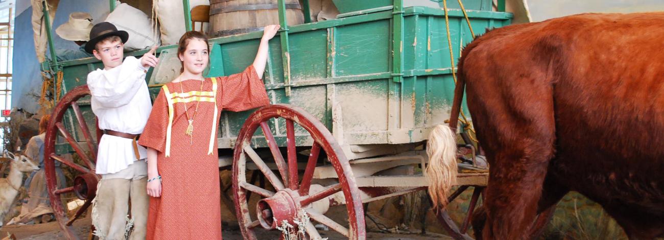 two children in old fashioned garb standing next to display of a covered wagon