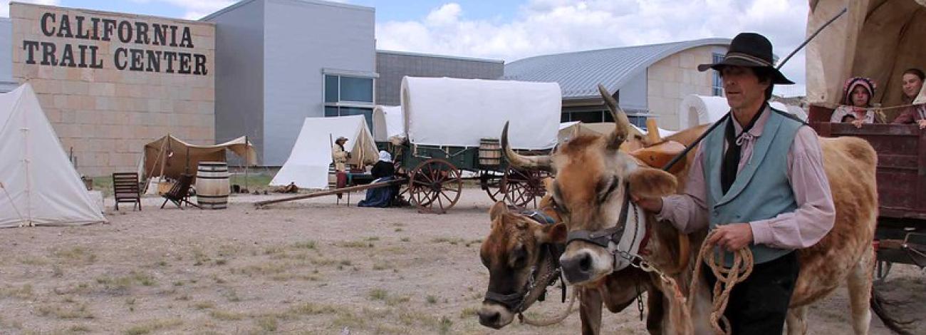 A person dressed in a hat and boots leads cattle pulling a wagon in front of a building that says California Trail Center.
