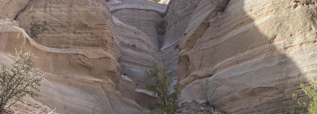Two hikers enjoy a hike within the Slot Canyon. Banded hoodoos tower on both sides of the trail with a bright blue sky.