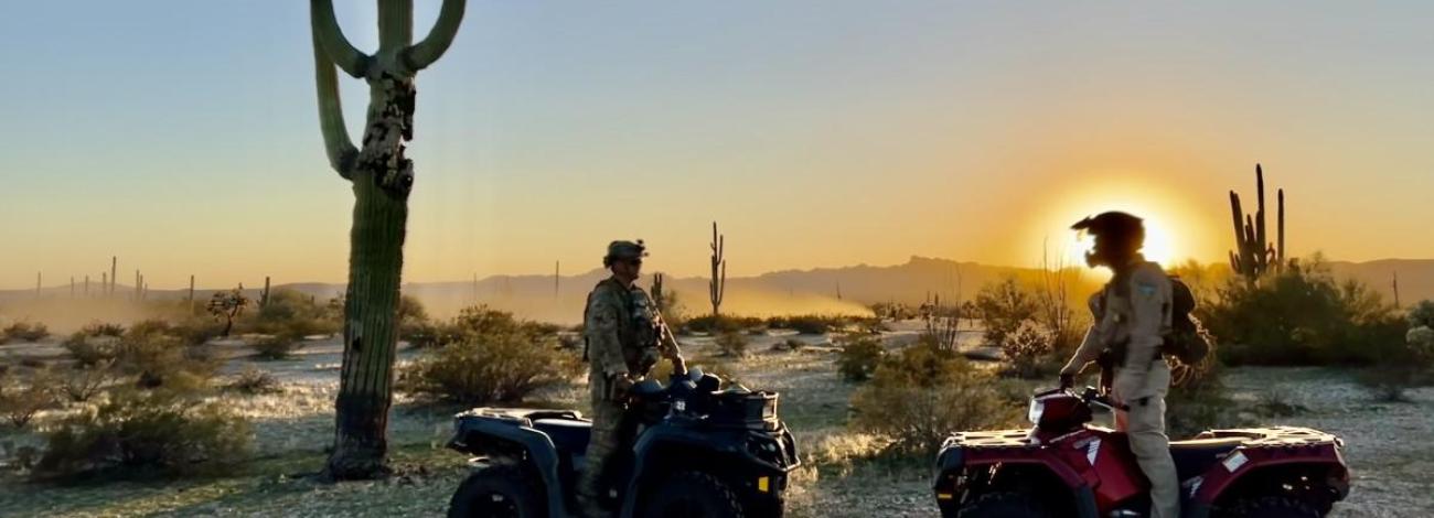 Two BLM rangers astride ATVs talk on a desert landscape with the sun in the background.