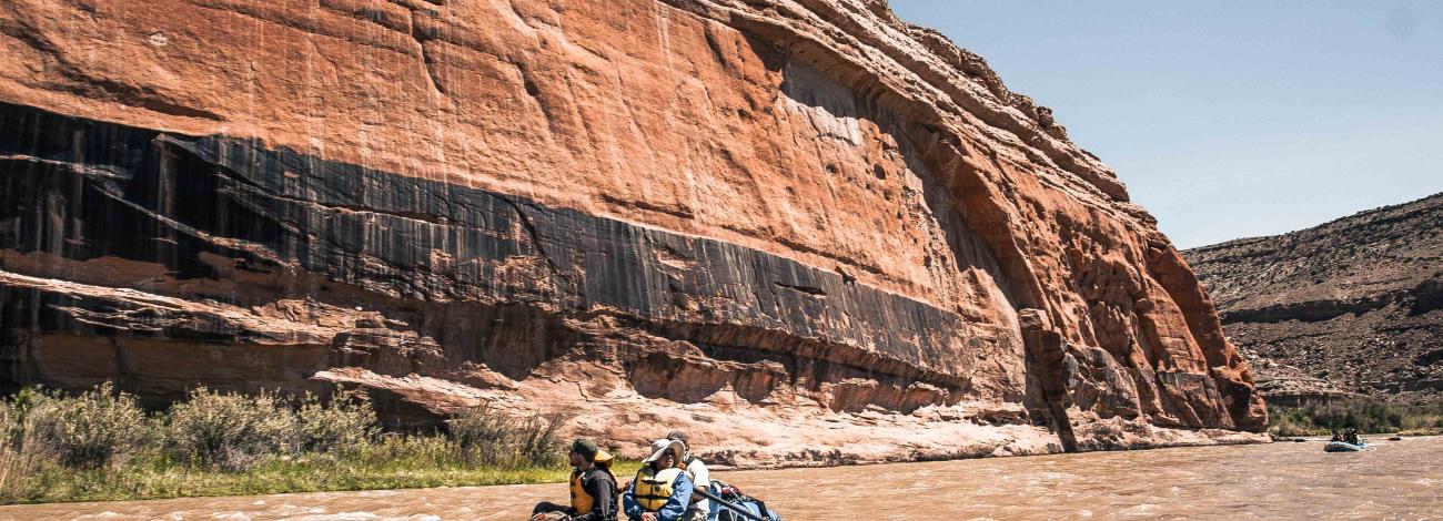 a blue raft with people in it goes down a muddy river with a red and black canyon wall in the background