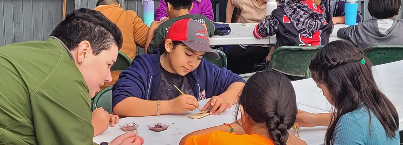 Photo. Uniformed Ranger leans over a table assisting students with a paint making activity. Students hold brushes and are painting on tan colored rocks. Ten students can be seen in the image, sitting around tables in the daytime. 