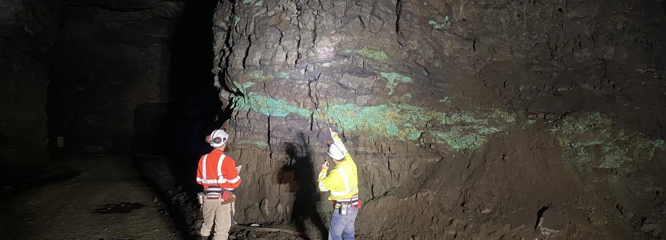 Two miners wearing safety gear inspect a rocky underground wall with green mineral deposits illuminated by a flashlight.