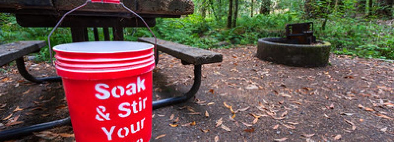 A bucket hanging from a picnic table and a fire pit in forested campsite.