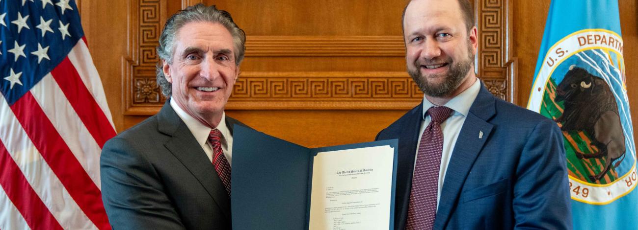 Two men in suits shake hands while holding a certificate in a formal wood-paneled room with U.S. flags behind them.