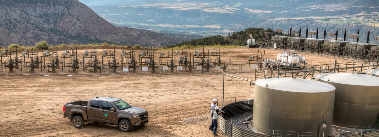 Two workers in hard hats inspect large industrial tanks at a remote oil or gas facility surrounded by mountainous terrain.