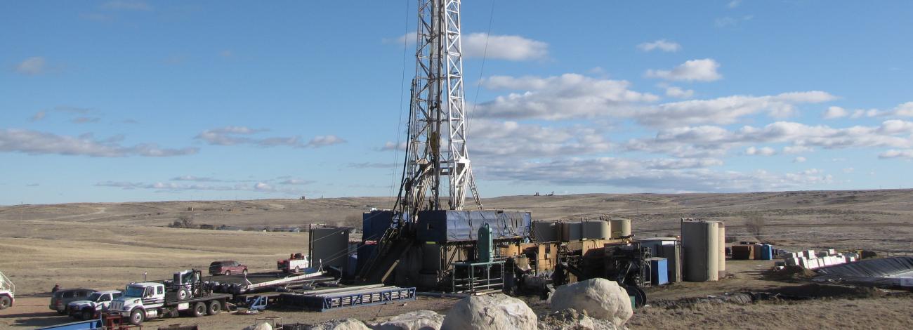 Oil drilling rig set against a clear blue sky with scattered clouds. The foreground features large rocks and vehicles.