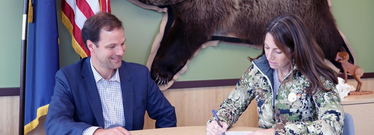 BLM AK State Director Kevin Pendergast and DOI Deputy Secretary Kate MacGregor sit at a table in front of the Alaska and American flags and a hung bear pelt signing a piece of paper.