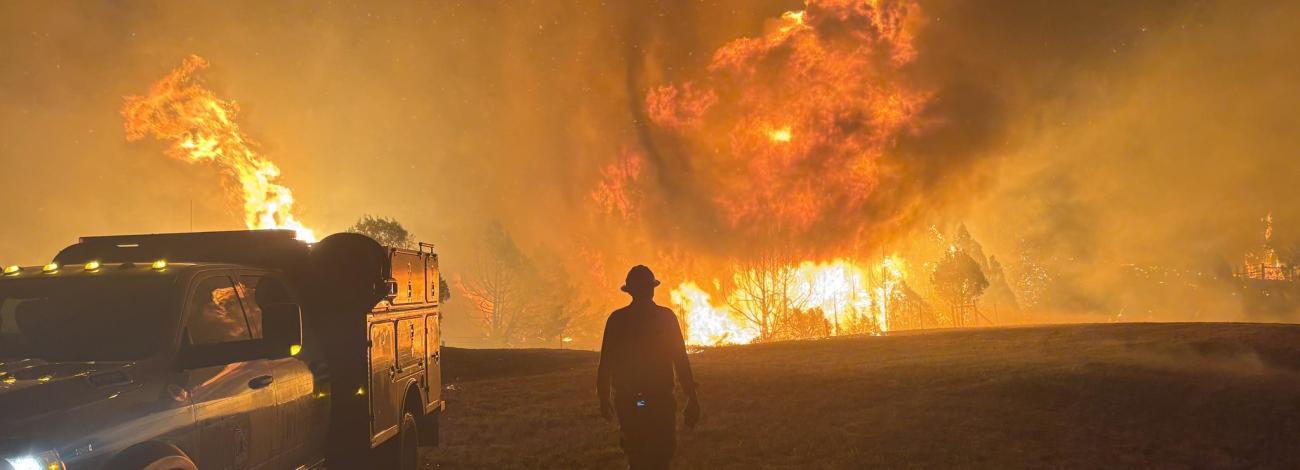 A wildland firefighter is silhouetted by a wildfire in the background as they walk near a truck.