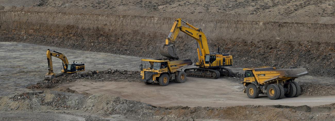 An excavator loads material into a large dump truck as another dump truck waits to be loaded.