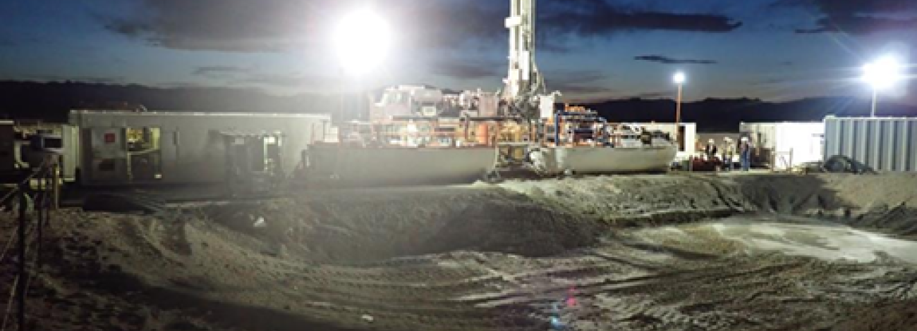 A white oil drill stands above several office trailers above an excavated pit about 50 meters wide and 5 meters deep. It is night. The scene is lit by 3 industrial light towers.