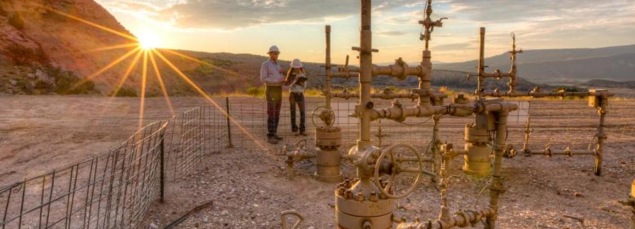 Two BLM employees work near oil and gas pipes with a mountain and the setting sun in the background.