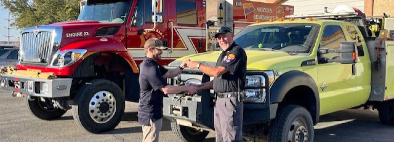 Two men exchange keys in front of fire engines.