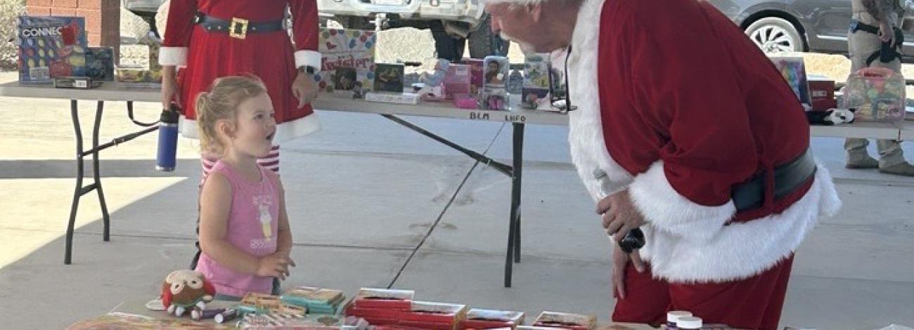 Mrs. Claus standing in the background watching a little girl visit with Santa Claus next to the table where children chose a present while visiting.