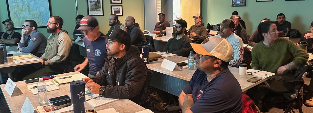 A room full of trainees sitting at tables observing a presentation.