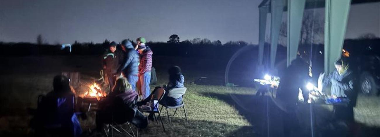 A group of people gathered around a campfire at night, with some sitting in chairs and others preparing food under a shelter.