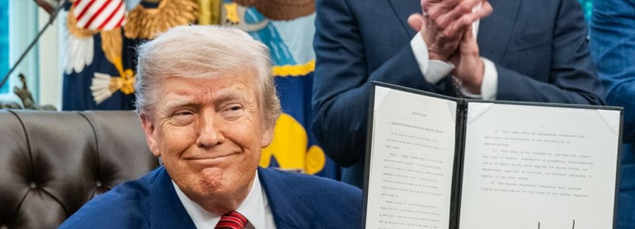 President Trump sits at his desk and holds up one of the energy executive orders he signed with Secretary of the Interior Burgum standing behind him clapping in the oval office.