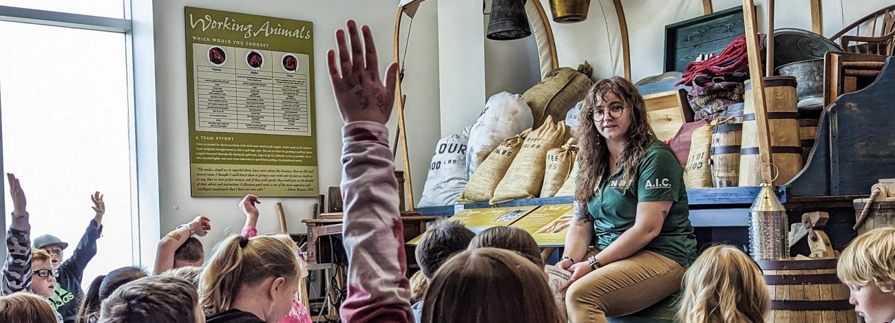 Students raise hands during education program with ranger sitting in front of replica covered wagon